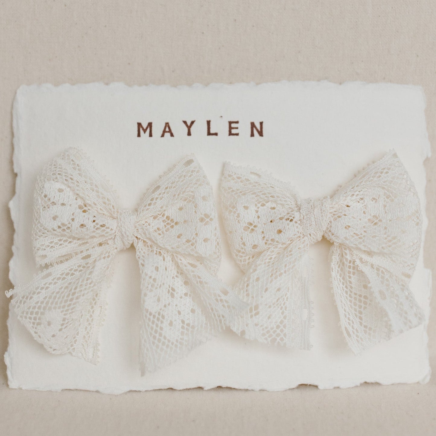 Two lace bows on a white card with 'MAYLEN' printed on it against a beige background