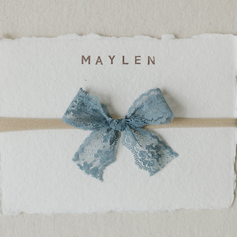 Blue bow headband on a textured white card with 'Maylen' printed on it, against a beige background.