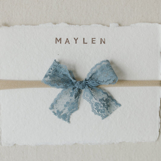 Blue bow headband on a textured white card with 'Maylen' printed on it, against a beige background.