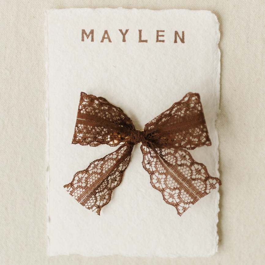 Brown lace bow on a white card with 'MAYLEN' branding on a beige background