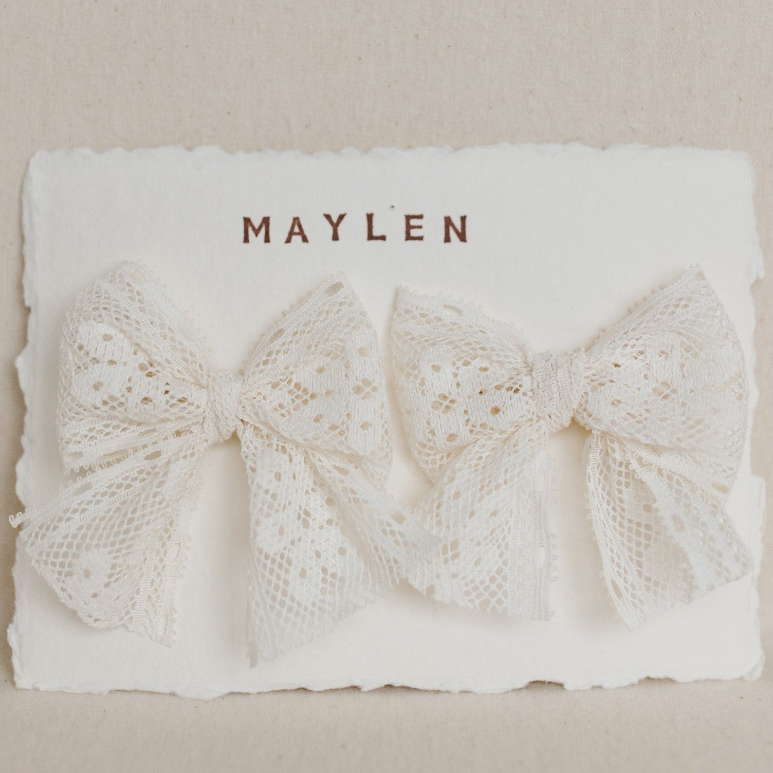 Two lace bows on a white card with 'MAYLEN' printed on it against a beige background