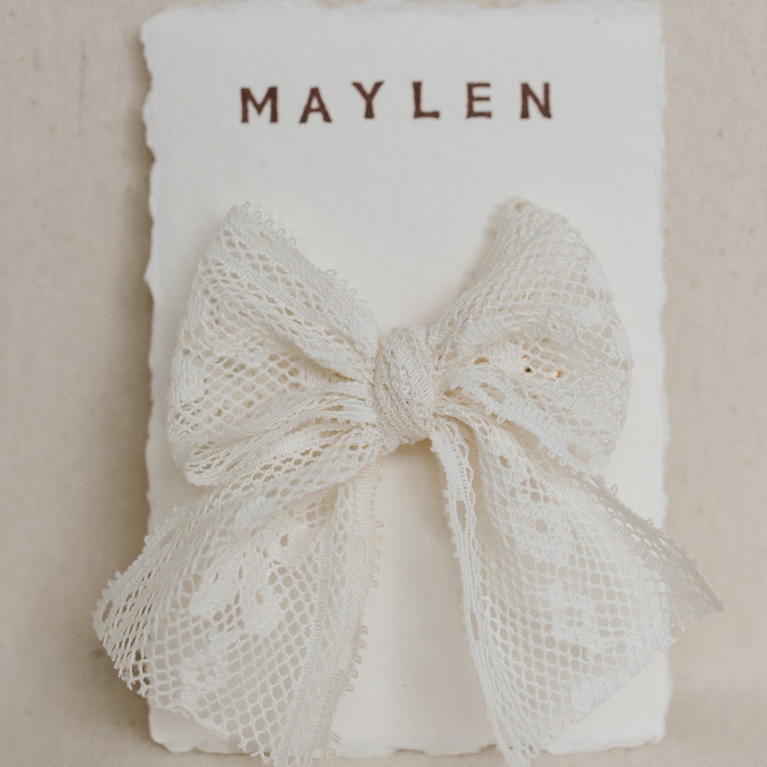 White lace bow on a beige background with 'MAYLEN' text.