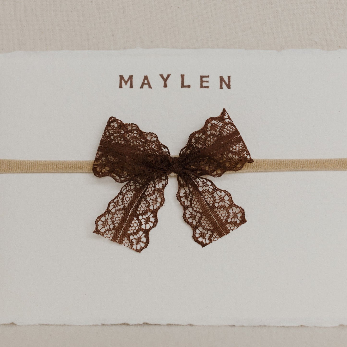 White card with a brown lace bow and 'MAYLEN' text on a beige background