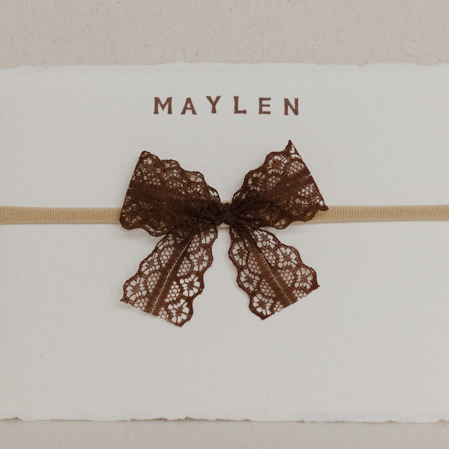 White card with a brown lace bow and 'MAYLEN' text on a beige background